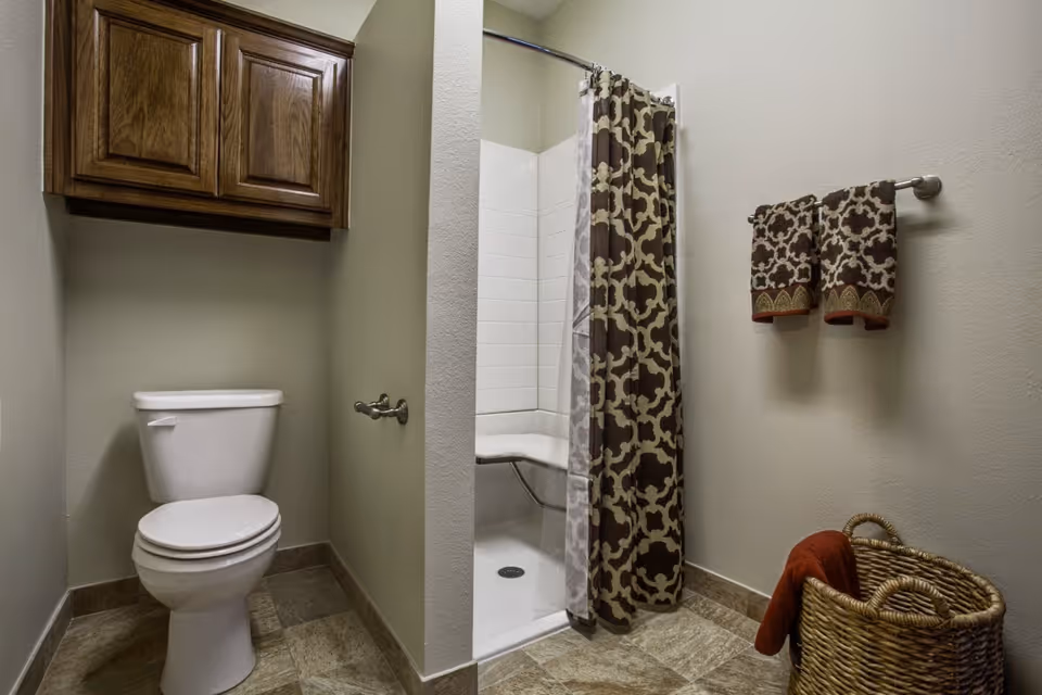A bathroom with a white toilet, a wooden cabinet mounted on the wall above the toilet, a walk-in shower with a patterned shower curtain, two matching patterned towels hanging on a towel rack, and a wicker basket with a red towel inside on the floor.