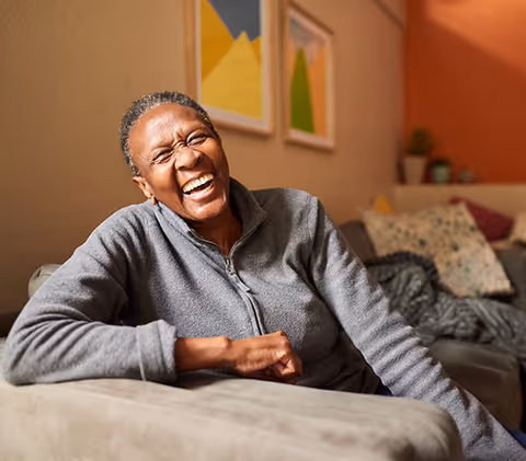 An elderly person with short gray hair wearing a gray zip-up jacket is sitting on a couch, smiling and laughing joyfully. Behind them, there are colorful abstract paintings on the wall and some cushions on the couch.