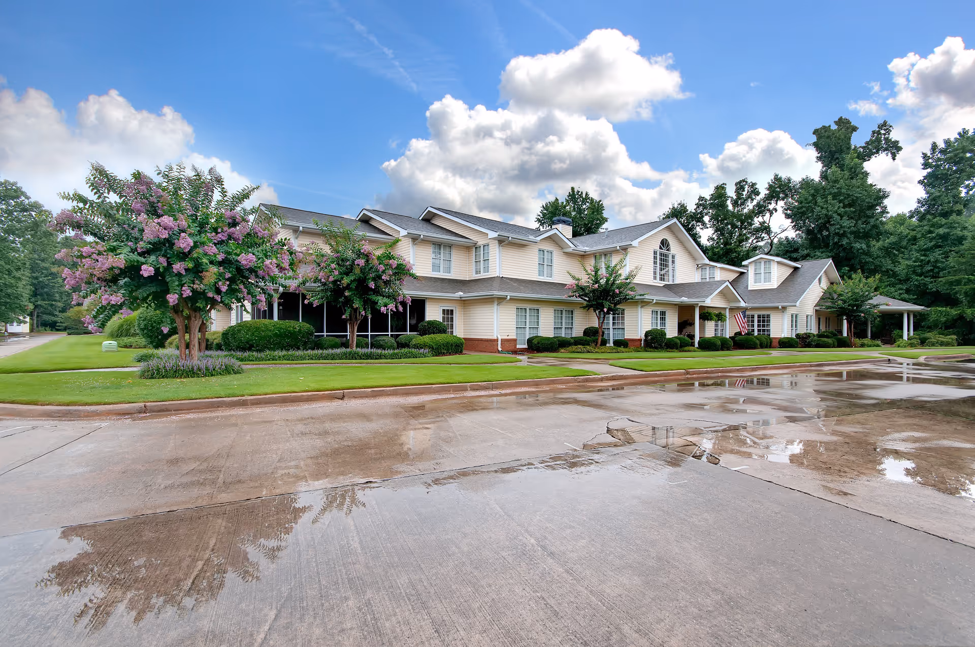 Exterior view of a two-story senior living facility building with beige siding, multiple windows, and a covered entrance. The building is surrounded by well-maintained green lawns, flowering trees, and shrubs. The sky is partly cloudy with blue patches visible. The concrete driveway in front of the building is wet, reflecting parts of the building and trees.