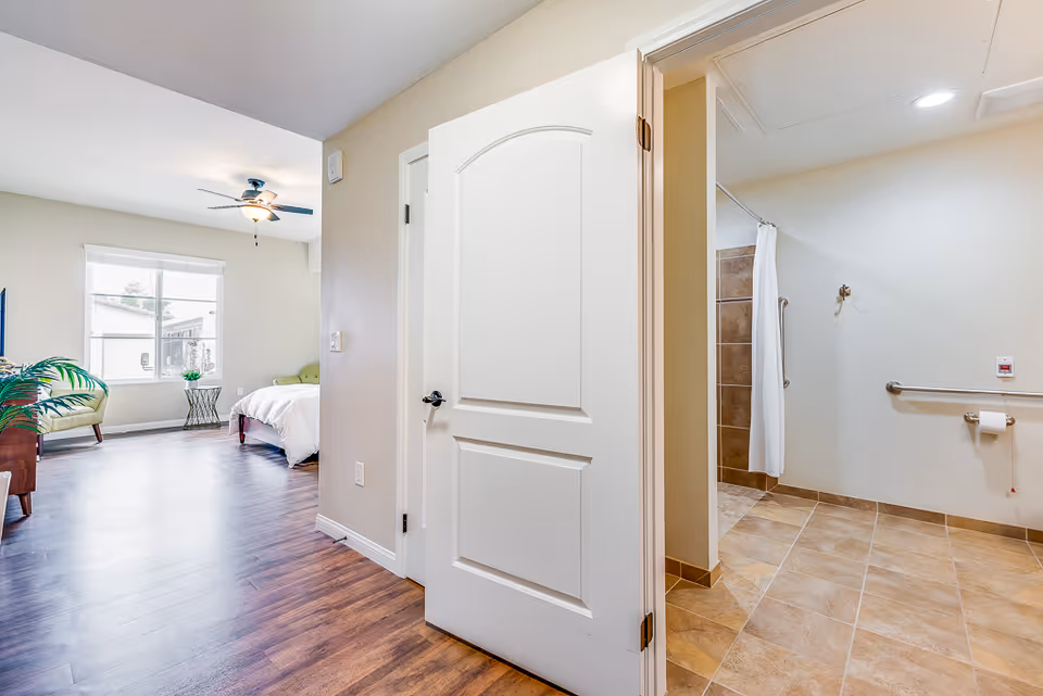 View of a senior living facility room showing a bedroom area with a bed, chair, and window with blinds, and an adjacent bathroom with tiled floor, grab bars, a shower area with a curtain, and a toilet paper holder.
