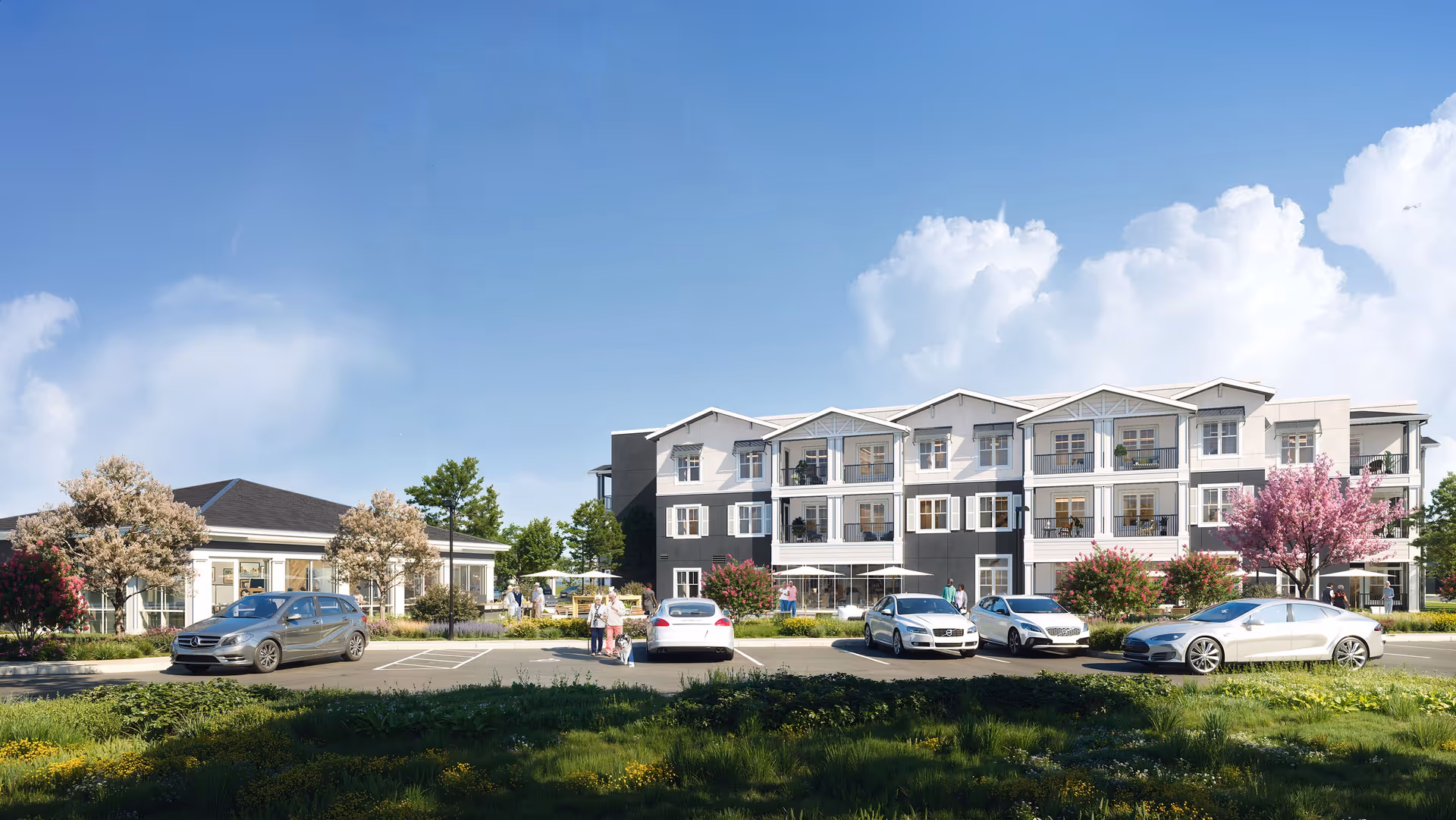 Front exterior of a three-story senior living building with landscaped grounds, parked cars and a few people under a clear blue sky.