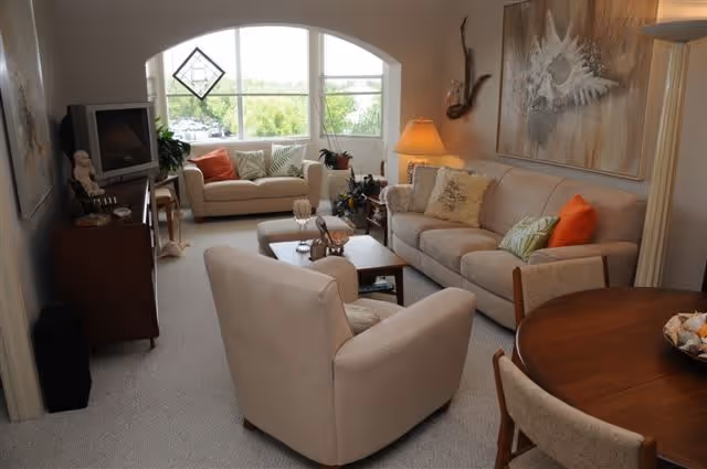 A cozy living room with beige sofas and armchairs arranged around a wooden coffee table. There are decorative pillows on the sofas, a lamp on a side table, and a large window with an arched top letting in natural light. A TV sits on a wooden stand to the left, and a round wooden dining table with chairs is partially visible on the right. The walls are decorated with artwork and a mounted decorative piece.