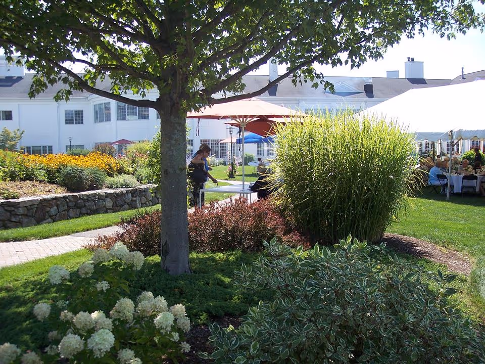 A landscaped garden area with various plants, flowers, and a tree in the foreground. In the background, there are people seated under a large white tent and colorful umbrellas near a white building, suggesting an outdoor event or gathering at a senior living community.