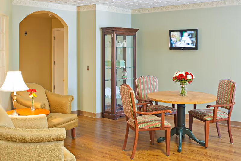 A cozy sitting area in a senior living facility with two beige armchairs around a small wooden side table with a lamp and a small vase of flowers. Nearby is a round wooden table with four striped upholstered chairs and a vase of red and white flowers in the center. A wooden display cabinet with glass doors holds decorative items, and a flat-screen TV is mounted on the light green wall.