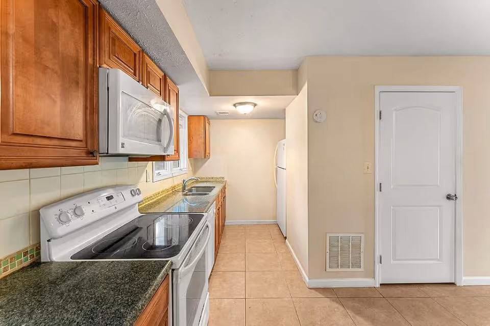 A narrow kitchen with wooden cabinets, a white electric stove with a glass cooktop, a white microwave above the stove, a double sink with a window above it, beige tiled floor, and a white refrigerator at the far end. There is a white door on the right wall and a ceiling light fixture near the refrigerator.