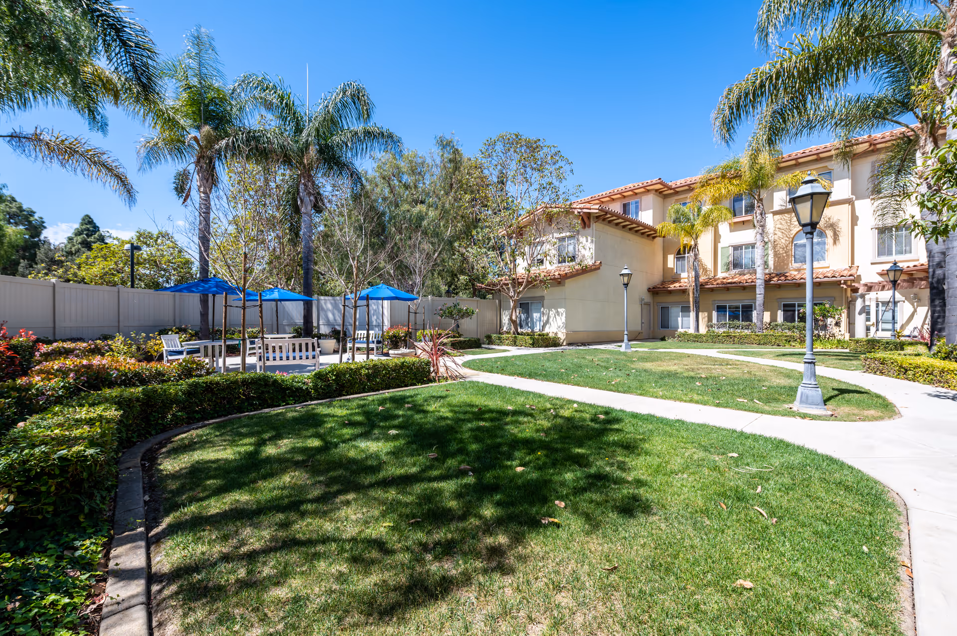 Outdoor garden area at Camarillo Senior Living with green grass, palm trees, and a paved walkway. There are benches and tables with blue umbrellas for shade, surrounded by various plants and flowers. The senior living building is visible in the background under a clear blue sky.
