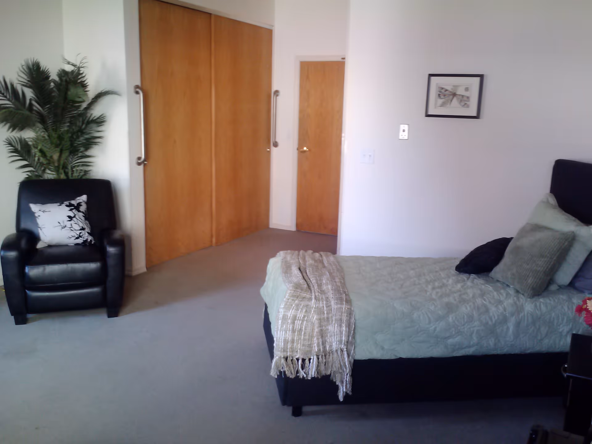 A bedroom with a single bed covered in light green bedding and multiple pillows. There is a beige throw blanket draped over the foot of the bed. To the left, there is a black armchair with a decorative pillow and a tall green plant behind it. The room has light-colored walls, a framed picture on the wall, and wooden closet doors with metal handles.
