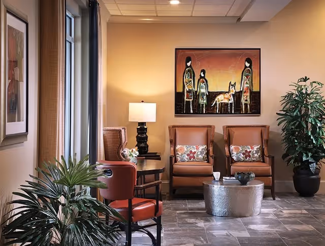 A cozy sitting area with two brown armchairs featuring floral cushions, a round metallic coffee table with a cup and decorative items, a black table lamp on a side table, a red chair, potted plants, and a colorful abstract painting of three figures and a dog on the wall.