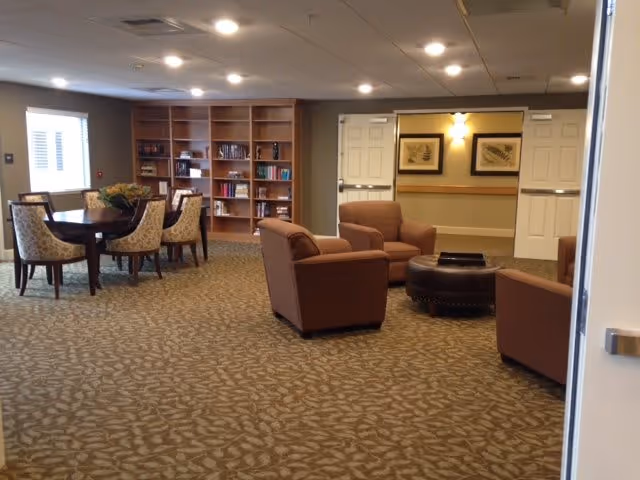 A spacious common area in an assisted living facility featuring a round table with four upholstered chairs on the left, a set of three brown armchairs arranged around a round ottoman in the center, and a large wooden bookshelf filled with books against the back wall. The room has patterned carpet, recessed ceiling lights, and double doors leading to a hallway with framed artwork.