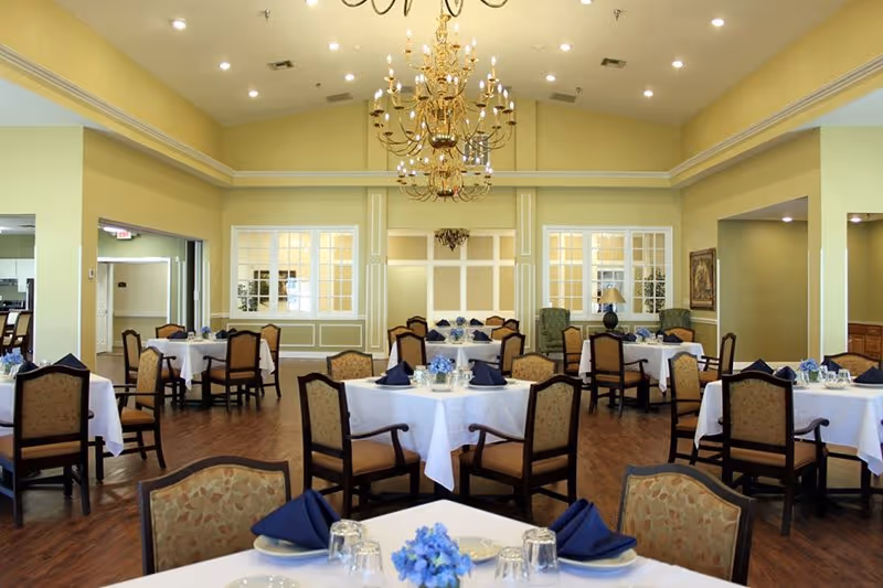 A spacious dining room in an assisted living facility with multiple round tables covered with white tablecloths, each set with blue napkins, glassware, and small blue floral centerpieces. The room features wooden chairs with cushioned seats, a polished wooden floor, cream-colored walls, large windows, and an elegant gold chandelier hanging from the ceiling.
