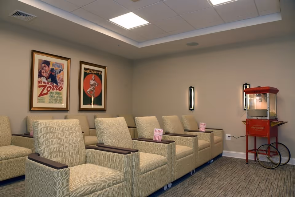 A cozy movie theater room with two rows of beige patterned armchairs, each equipped with a small wooden armrest. On the right side of the room, there is a vintage-style red popcorn machine. The walls are decorated with framed movie posters, including The Mark of Zorro and The Wizard of Oz. The room has soft lighting from wall sconces and a ceiling with recessed lights.
