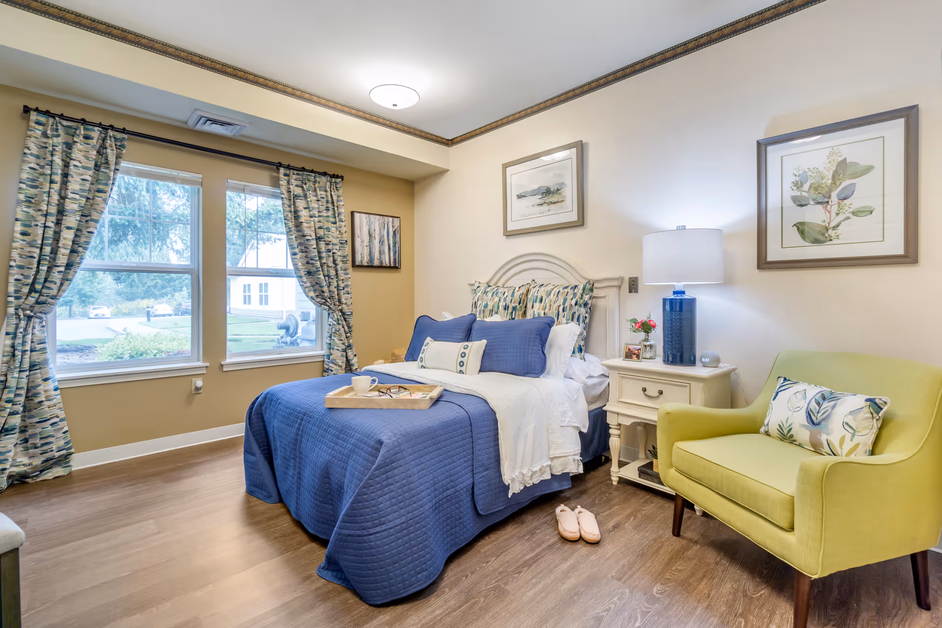 Bright furnished bedroom with a blue quilted bed, patterned curtains, a green armchair, and a bedside table with a lamp.