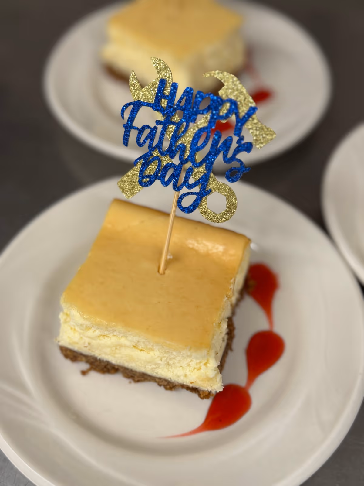 A slice of cheesecake on a white plate with a decorative toothpick topper that says 'Happy Father's Day' in blue and gold glitter. There is a red sauce decoration on the plate next to the cheesecake.