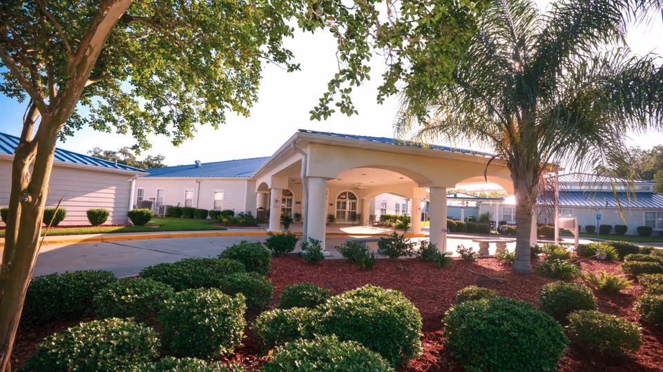 Exterior view of Riverbend Nursing & Rehabilitation Center showing a covered entrance with columns, surrounded by well-maintained landscaping including bushes, trees, and palm trees under a clear sky.
