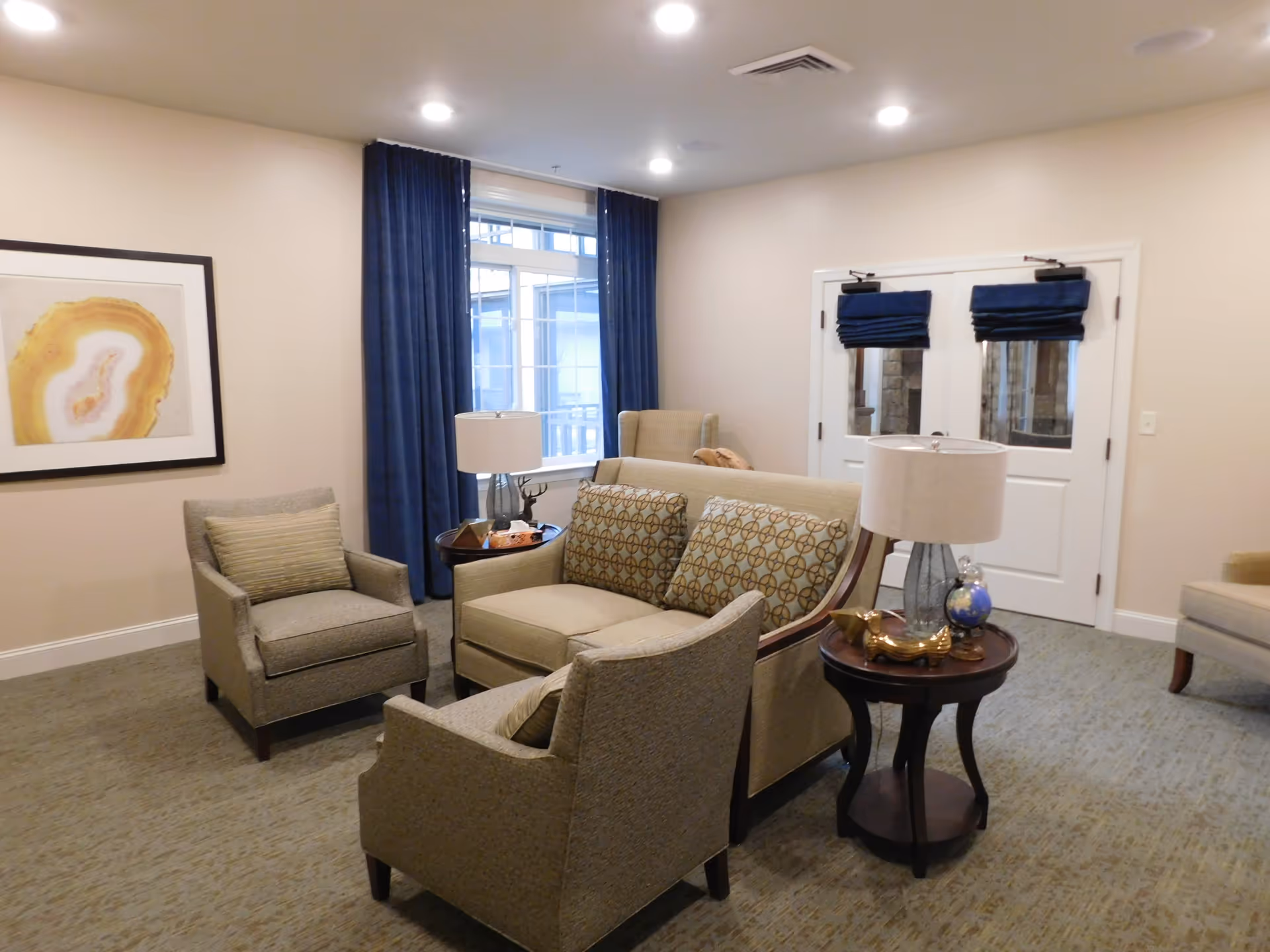 A cozy living room area with beige walls and carpeted floor. The room features a loveseat with patterned cushions, two armchairs with striped pillows, a round wooden side table with a lamp and decorative items, and a window with dark blue curtains. There is a framed abstract artwork on the wall and double doors with blue Roman shades.