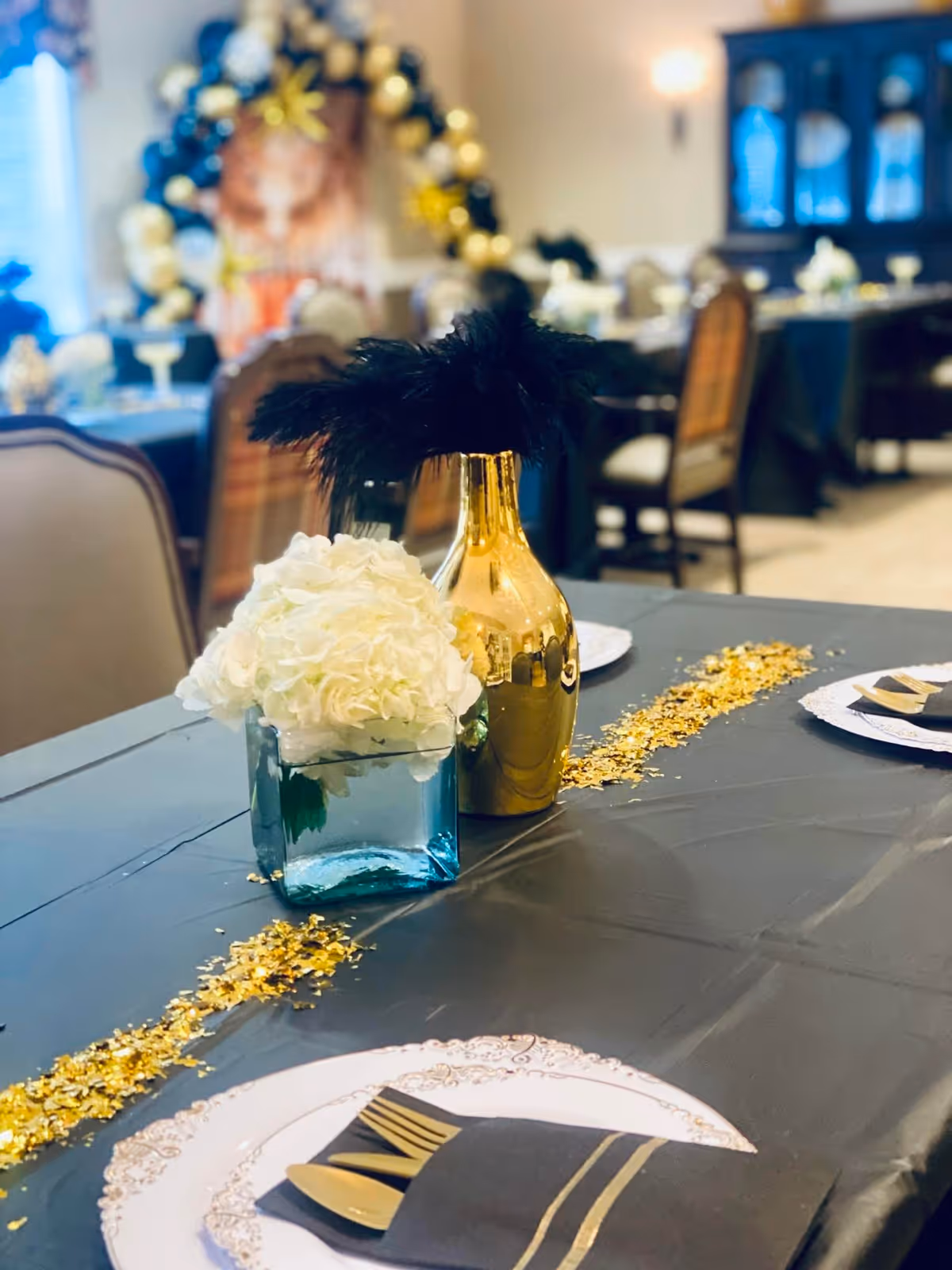 A dining table set for an event with a black tablecloth, gold and black cutlery wrapped in black napkins, a centerpiece featuring white flowers in a blue glass vase, and a gold vase with black feathers. The background shows more tables and chairs with similar decorations and a balloon arch in black and gold colors.