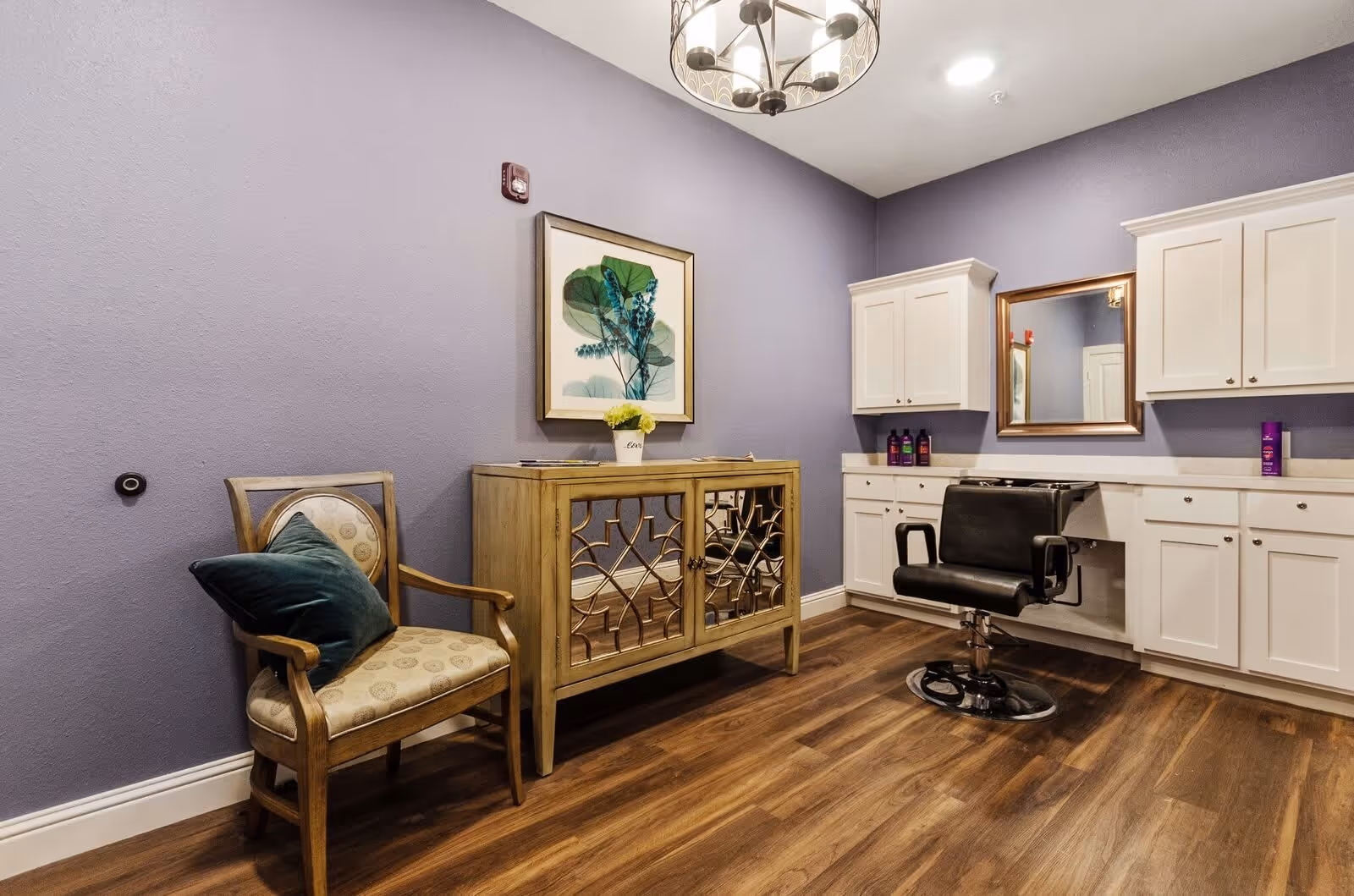 Interior room with purple walls and wood flooring featuring a wooden chair with a green cushion, a wooden cabinet with mirrored doors, white cabinets mounted on the wall, a black salon chair in front of a mirror, and a chandelier hanging from the ceiling.