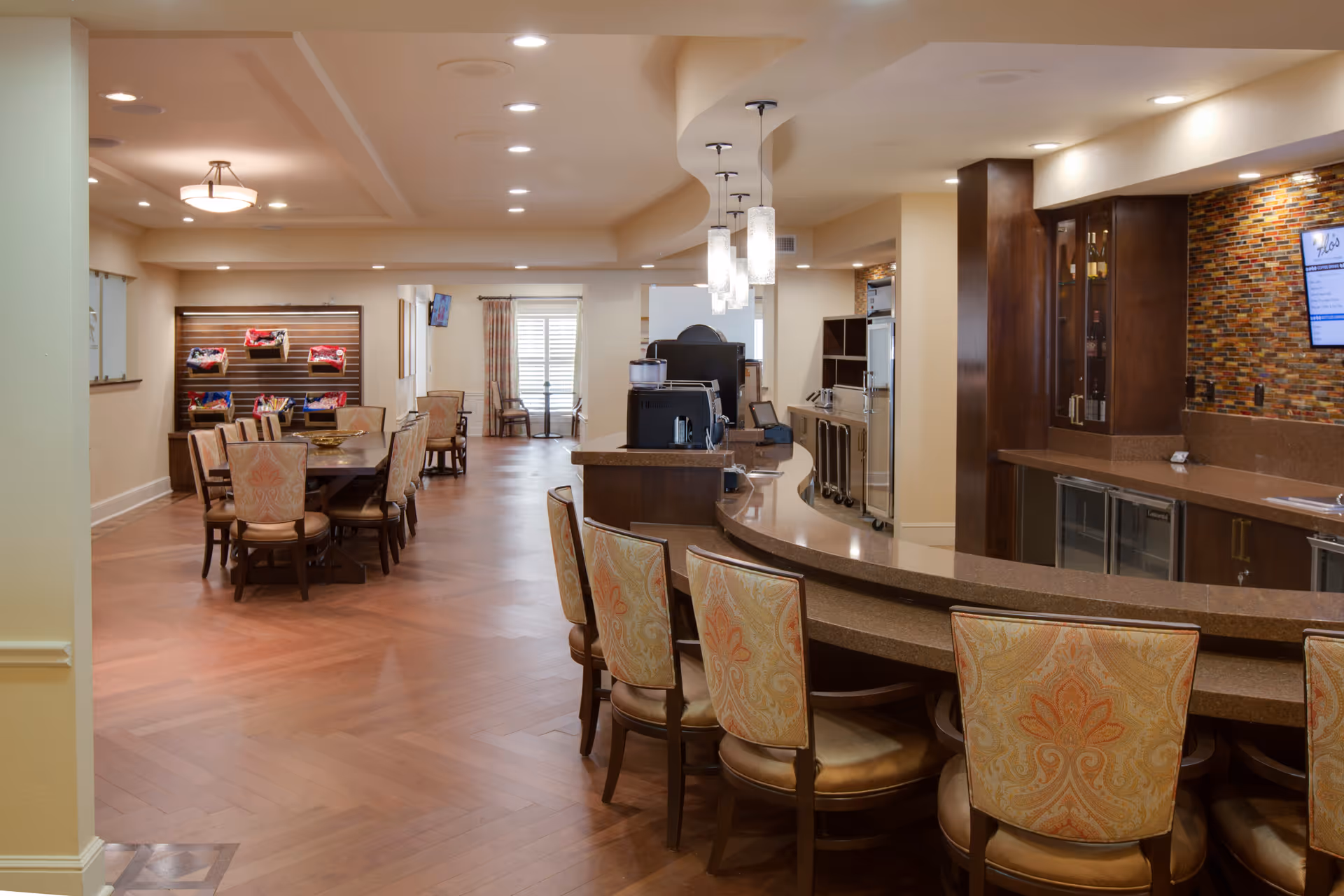 Interior view of a senior living facility dining and lounge area featuring a curved bar with upholstered chairs, a long dining table with chairs, wooden flooring, pendant lights, and a colorful tiled backsplash behind the bar area.
