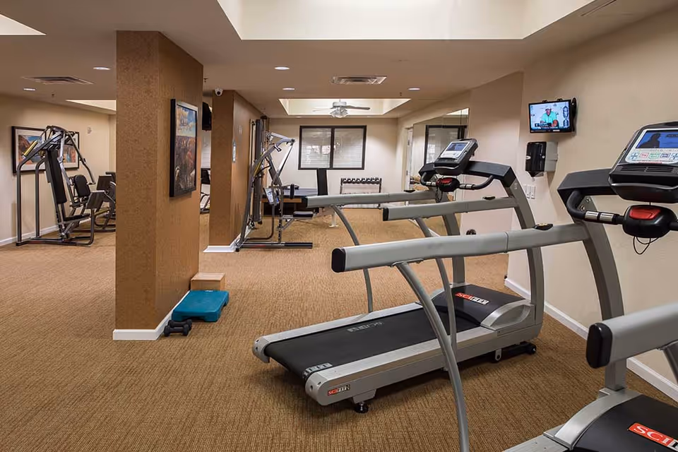Indoor fitness room with two treadmills in the foreground, weight machines along the walls, a set of dumbbells, and a step platform. The room has beige walls, carpeted floor, and a wall-mounted TV.