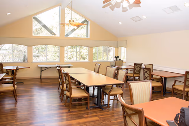 Well-lit dining room with multiple wooden tables and chairs, hardwood floors, large windows, and a vaulted ceiling with fans.