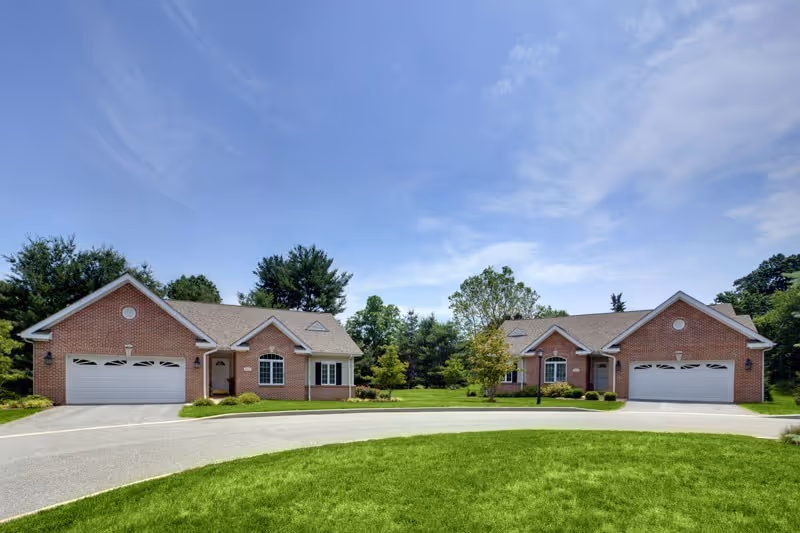 Two single-story brick houses with attached garages facing a curved driveway, surrounded by green grass and trees under a blue sky with some clouds.