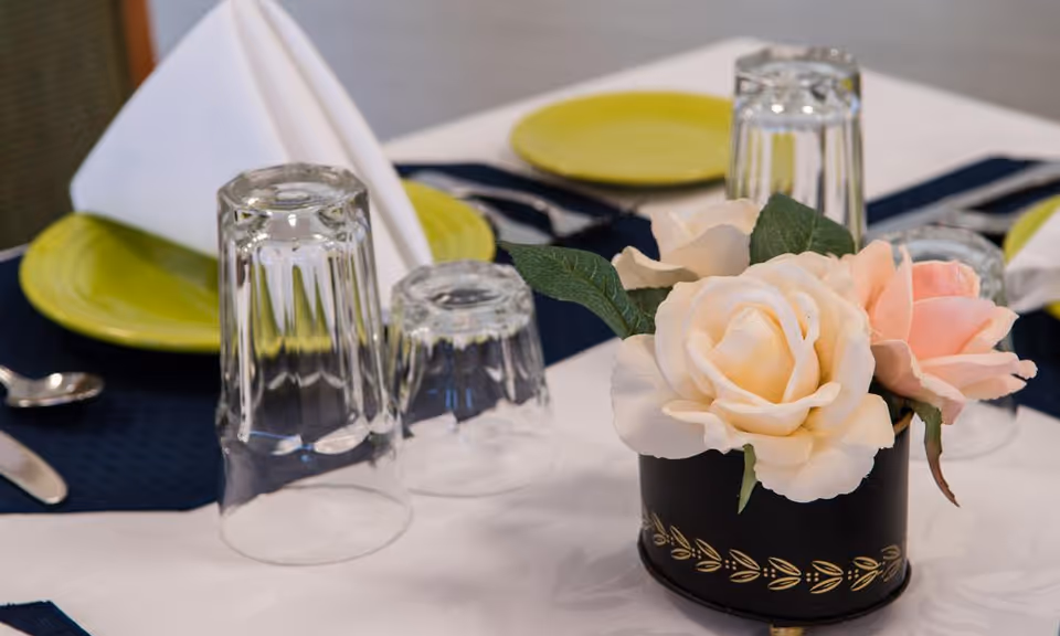 Close-up of a dining table set with upside-down clear drinking glasses, green plates, silverware, white folded napkins, and a small black vase holding cream and pink artificial roses on a white tablecloth with a dark blue placemat.