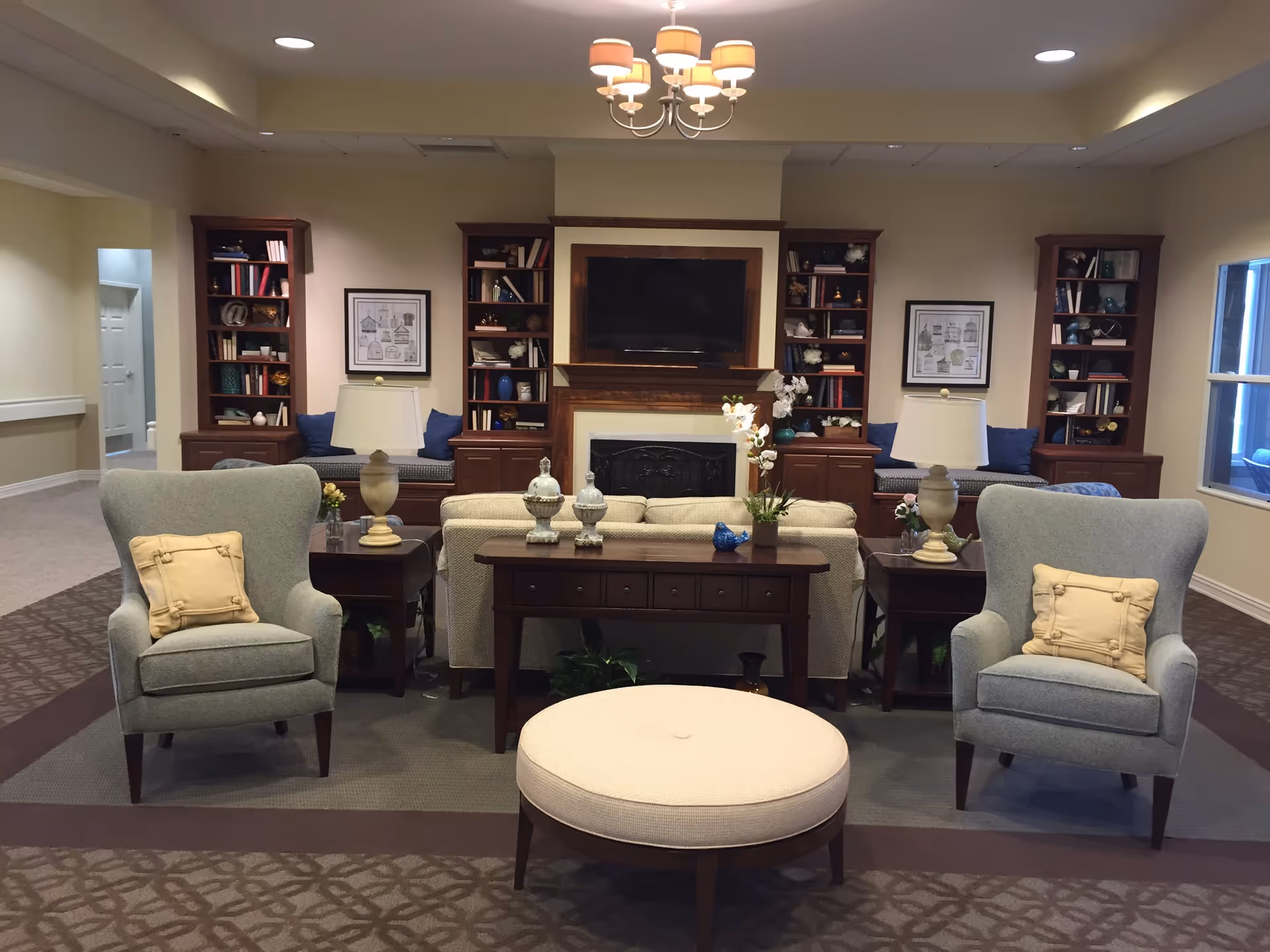 Communal lounge with two armchairs, ottoman, sofa, side tables and lamps facing a TV above a fireplace flanked by bookshelves.