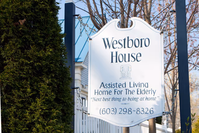 A white hanging sign for Westboro House, an assisted living home for the elderly, with the tagline 'Next best thing to being at home' and a phone number (603) 298-8326. The sign is mounted on a black metal frame with trees and a white fence in the background.