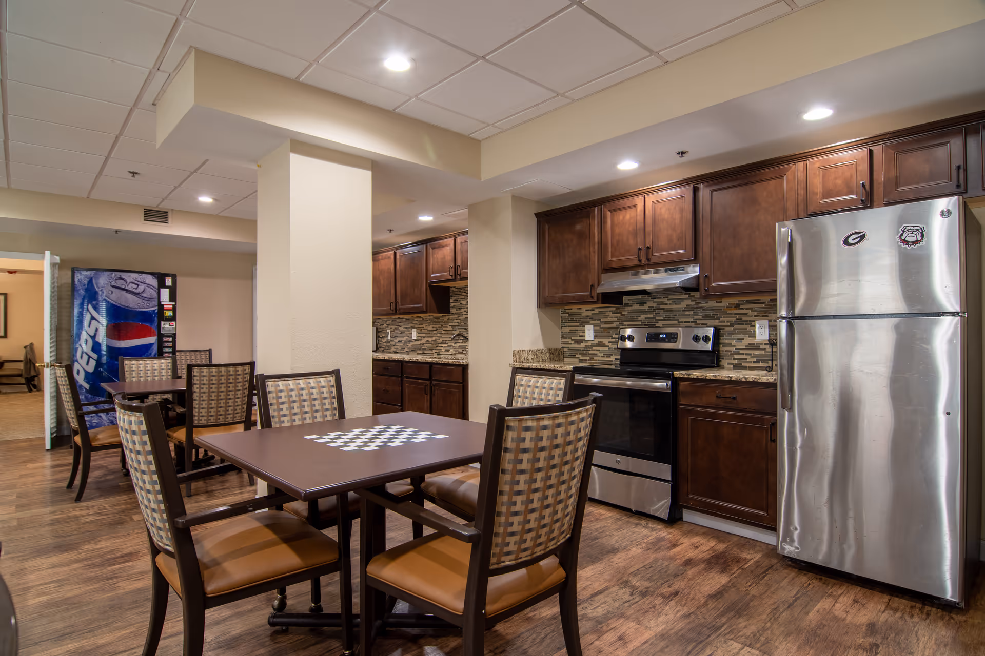 Interior view of a communal kitchen and dining area in a retirement community. The space features a stainless steel refrigerator and stove, dark wood cabinets, and a tiled backsplash. There are tables with checkerboard patterns on top and chairs arranged around them. In the background, a Pepsi vending machine is visible near an open doorway leading to another room.