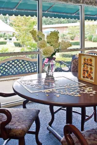 Round wooden table with puzzle pieces and a vase of flowers in a sunlit lounge.