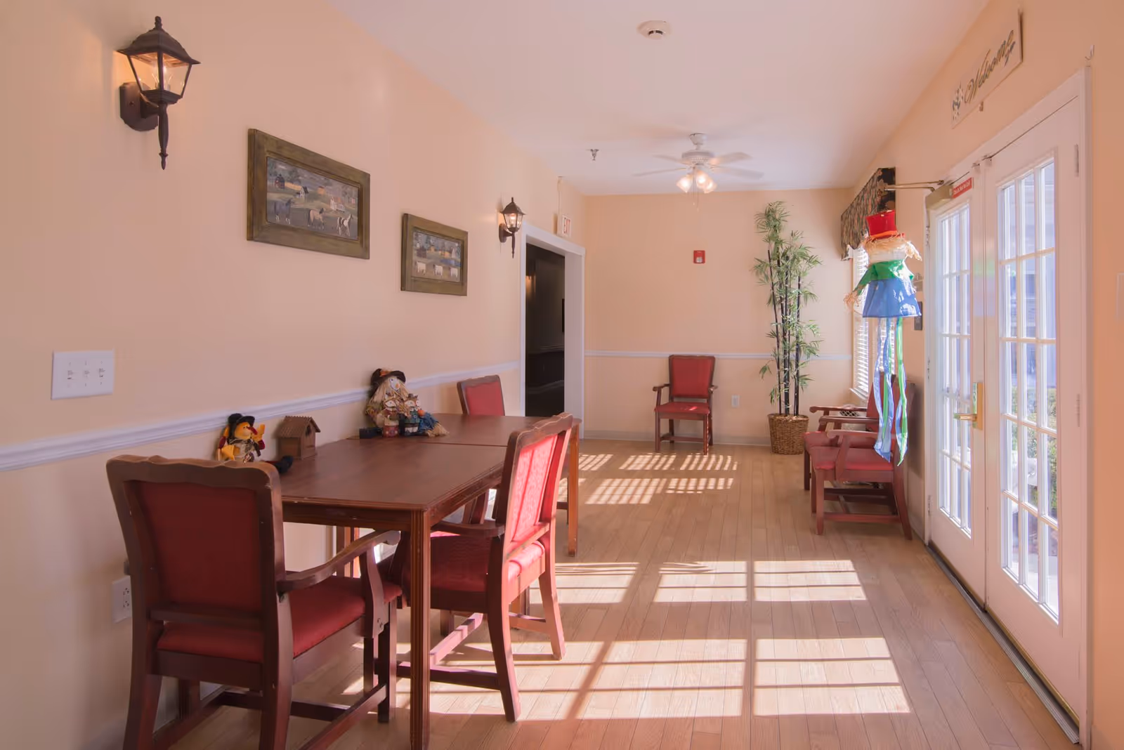 Sunlit communal dining area with a wooden table and red upholstered chairs, French doors, wall art and a potted plant.