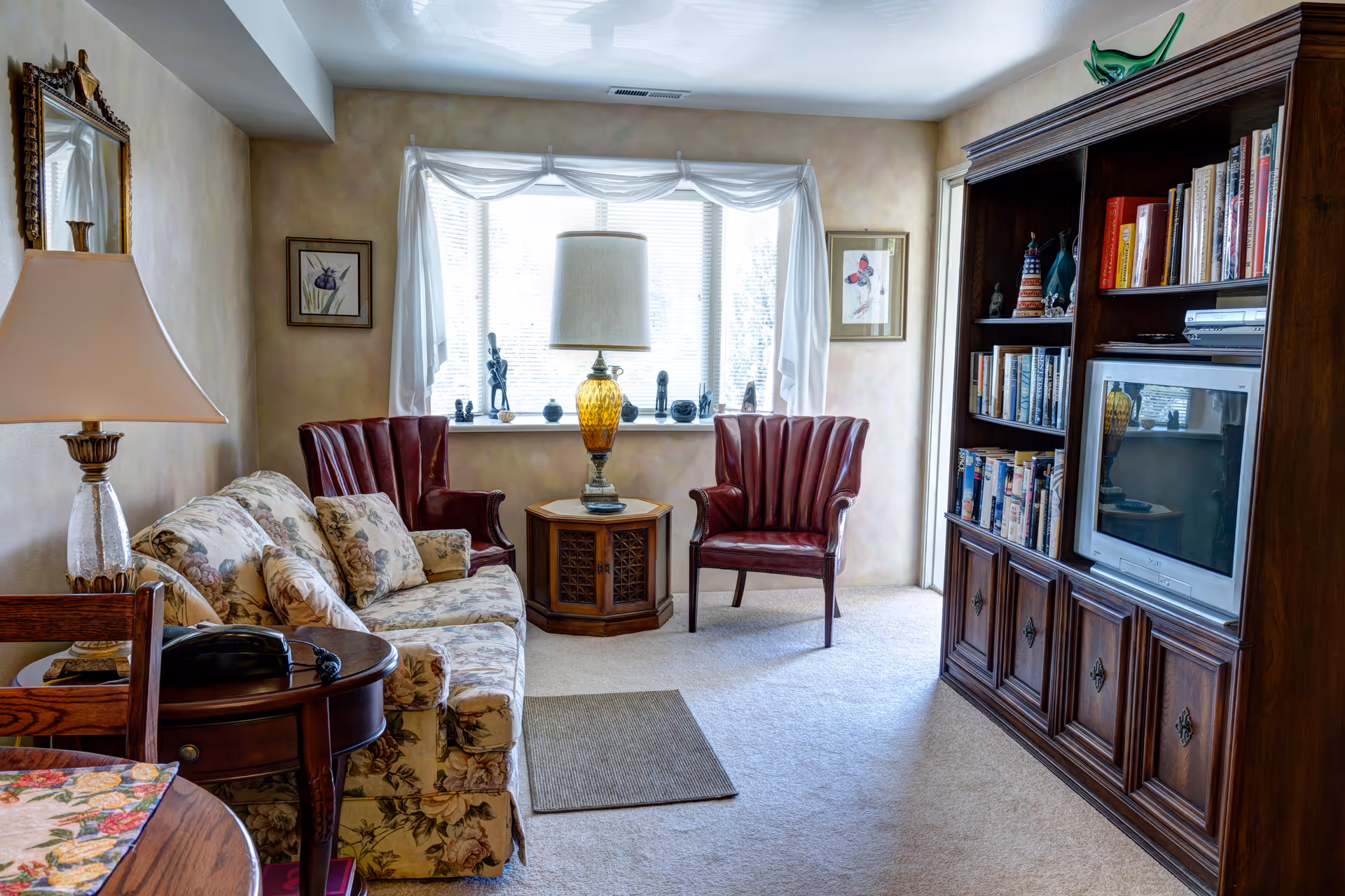 Cozy living room with a floral sofa, two red armchairs by a bright window, a central lamp on an end table, and a wooden entertainment center with a TV and books.