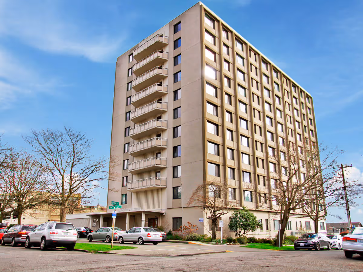 Exterior view of a multi-story senior living facility building named Hilltop, with several parked cars along the street and leafless trees around the property under a blue sky.