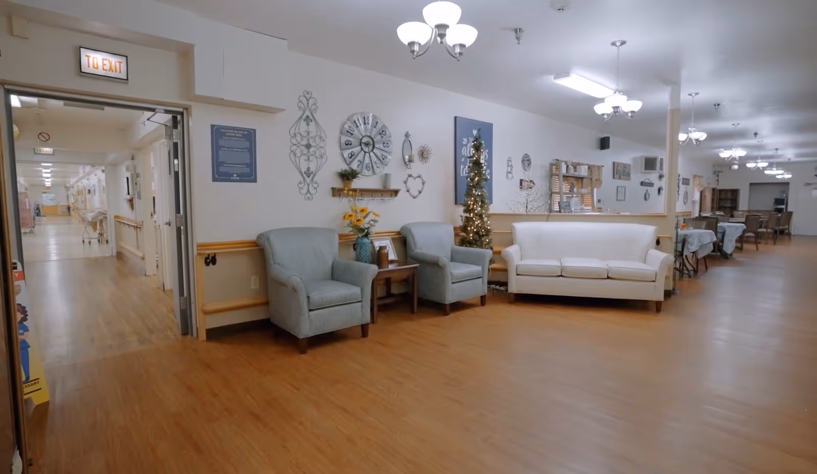 A spacious, well-lit common area in a senior living facility with wooden flooring, two light blue armchairs, a white sofa, and a small decorated Christmas tree. The walls are adorned with various decorative items including a large clock and framed art. In the background, there are tables and chairs arranged for dining or activities. An open doorway leads to a hallway with handrails and more rooms.