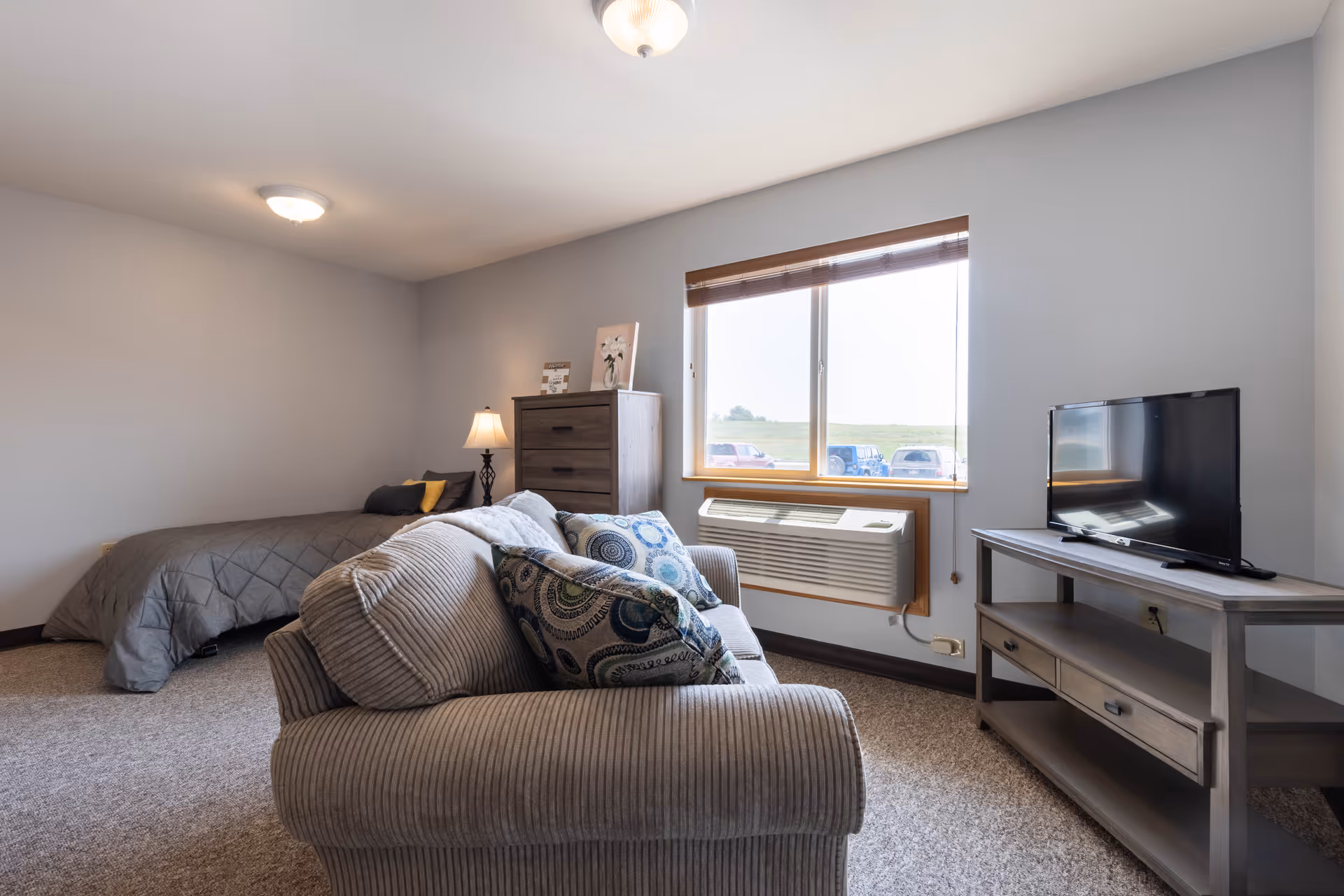 A cozy senior living bedroom featuring a bed with gray bedding, a wooden dresser with a lamp and decorative items, a window with a view of parked cars outside, a wall-mounted air conditioning unit below the window, a TV on a wooden stand, and a comfortable armchair with patterned cushions in the foreground.