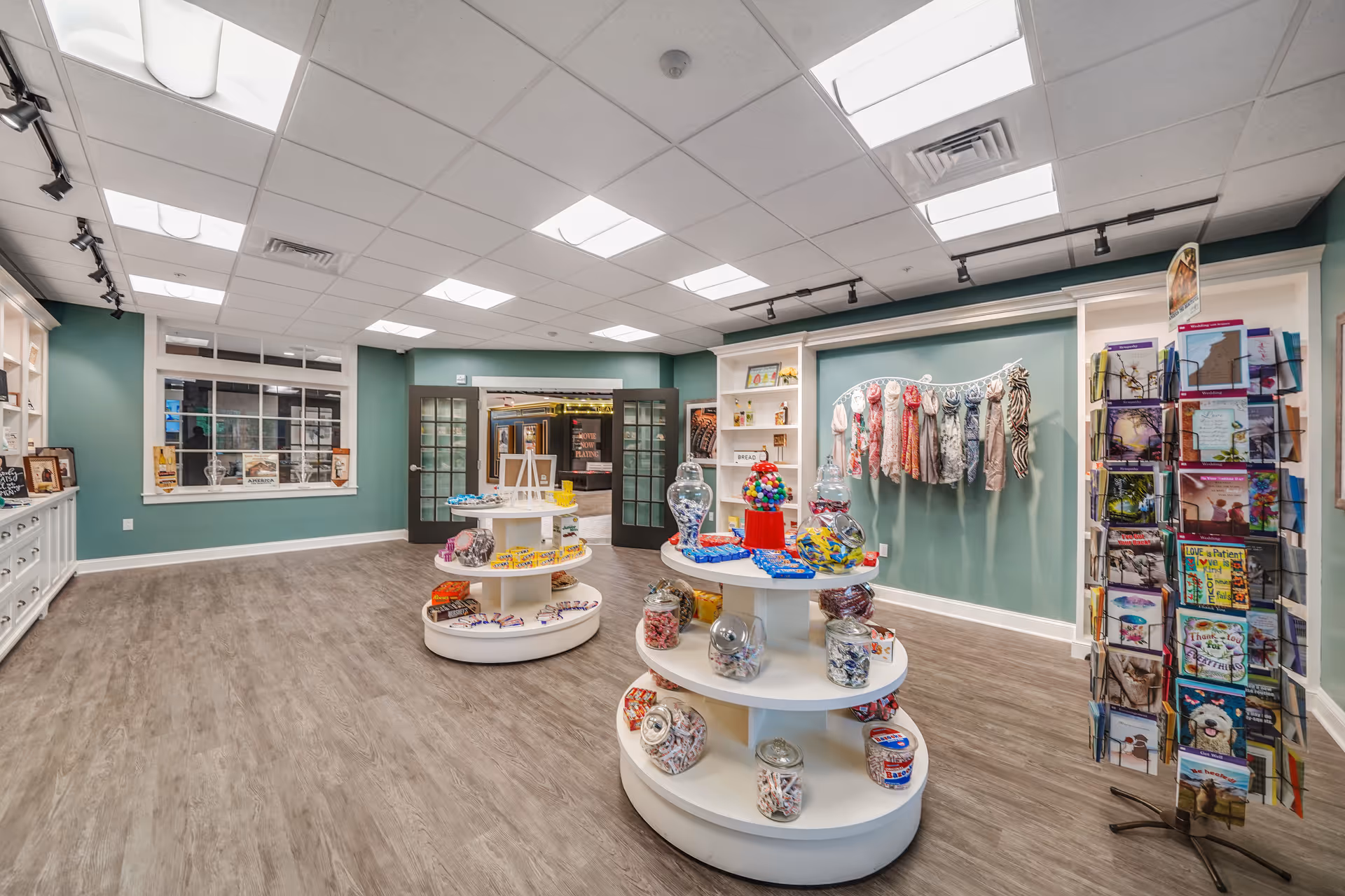 Interior view of a retail or gift shop area in a senior living facility with round display shelves holding jars of candy and snacks. There are scarves hanging on the wall and a rotating rack filled with greeting cards. The room has green walls, wood-look flooring, and a ceiling with recessed lighting. Double glass doors lead to another room in the background.