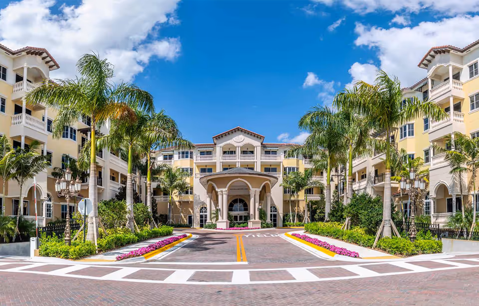 Front exterior entrance of a Mediterranean-style senior living building with a porte-cochère, palm trees, and a landscaped driveway under a blue sky.