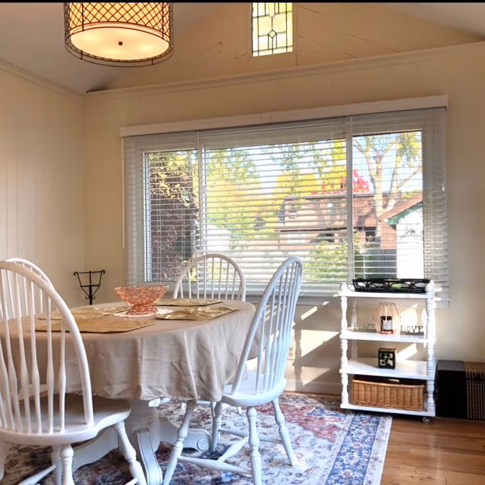 A bright dining room with a round table covered by a beige tablecloth, surrounded by four white wooden chairs. A decorative bowl is placed on the table. The room has a large window with white blinds, letting in natural light and showing a view of trees and houses outside. A white shelving unit with decorative items and baskets is positioned against the wall. A patterned rug covers the wooden floor, and a hanging light fixture is visible above the table.