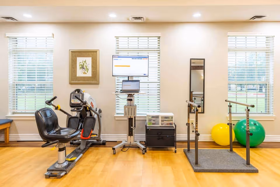 Well-lit rehabilitation room with a recumbent exercise bike, parallel bars, a computer workstation, and exercise balls in front of three windows.