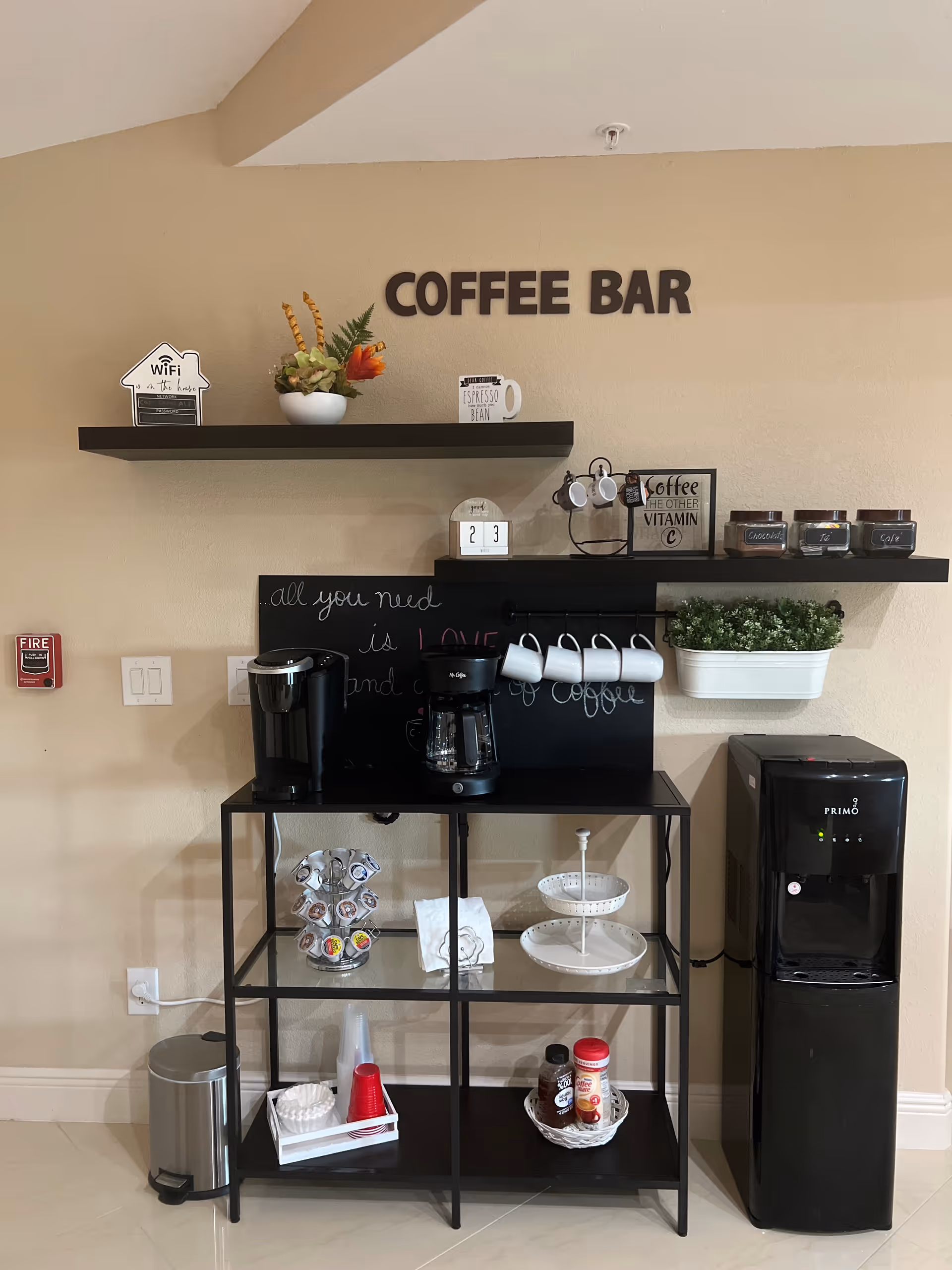 A coffee bar setup in an indoor space with beige walls. The setup includes two black wall shelves holding decorative items, coffee mugs, and labeled jars. Below the shelves is a black metal and glass stand with a coffee maker, a Keurig machine, coffee pods, disposable cups, and condiments. To the right of the stand is a black Primo water dispenser. A small trash can is on the floor to the left. The wall has the words 'COFFEE BAR' in large letters and a chalkboard with a partial quote about coffee.