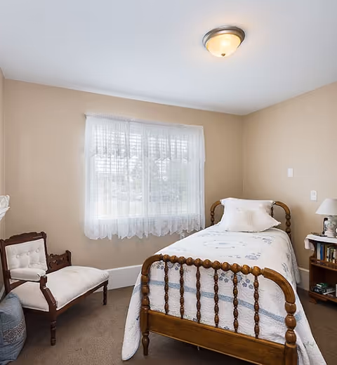 A small bedroom with beige walls and carpeted floor featuring a wooden single bed with a white and blue quilt and a white pillow. Next to the bed is a wooden nightstand with a lamp and books. A cushioned wooden chair is placed near a window with sheer white curtains, allowing natural light to brighten the room.