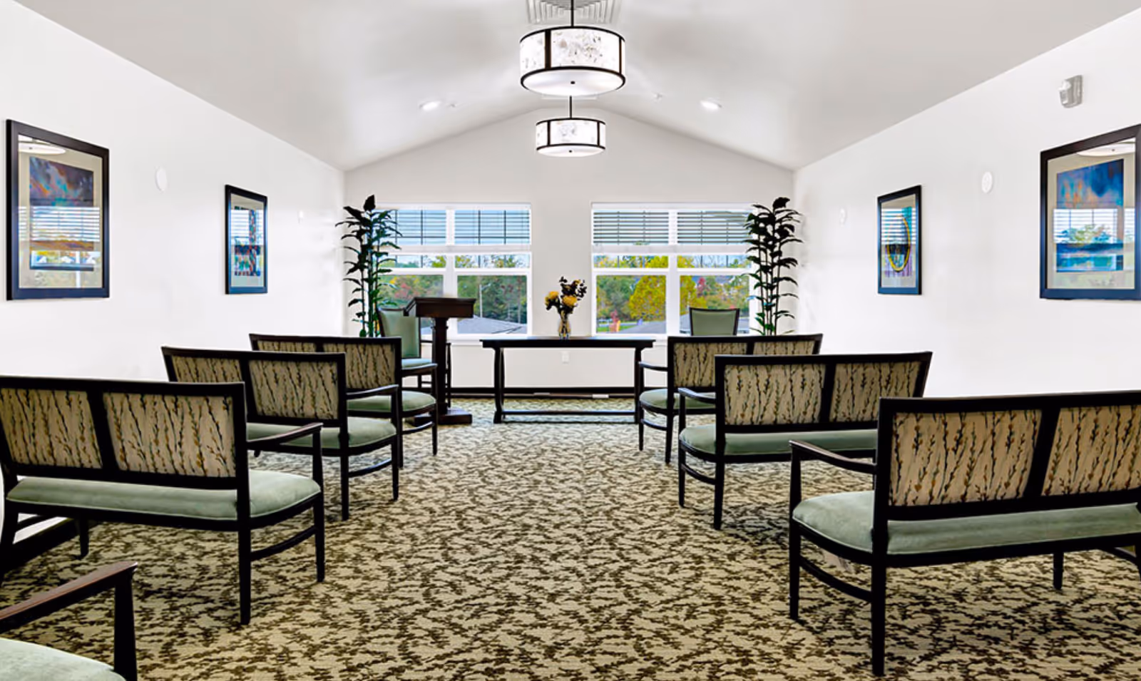 A bright, well-lit room with patterned carpet and rows of chairs facing a table and podium near large windows. The walls are white with framed artwork and tall plants on either side of the windows.