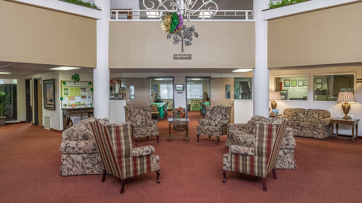 Open communal lounge with patterned armchairs and sofas arranged around a central table beneath a decorative chandelier and balcony.