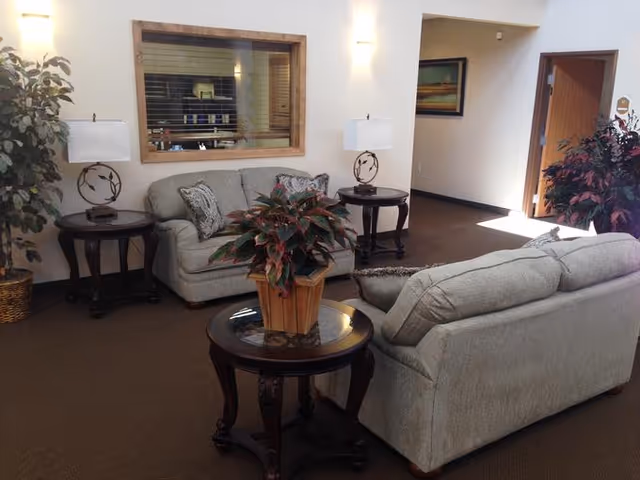 A cozy seating area in an assisted living facility with two gray upholstered sofas arranged around a round wooden coffee table holding a potted plant. Two matching wooden side tables with decorative lamps flank one sofa. A large window with wooden trim is set into the wall behind one sofa, and there are plants and framed artwork in the background.