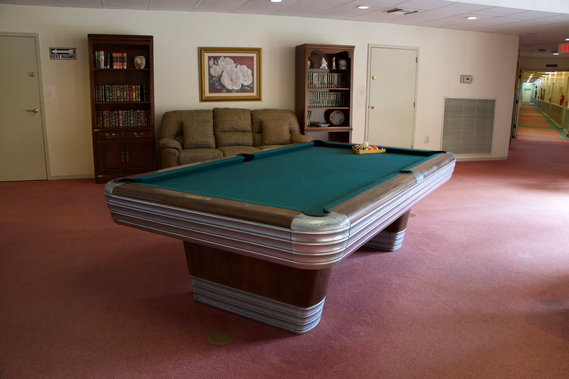A room with a green felt pool table in the center, a brown couch against the wall, two wooden bookshelves filled with books and decorative items, a framed floral painting above the couch, and a hallway visible to the right. The floor is carpeted in pink, and there is a sign on the left door indicating restrooms.