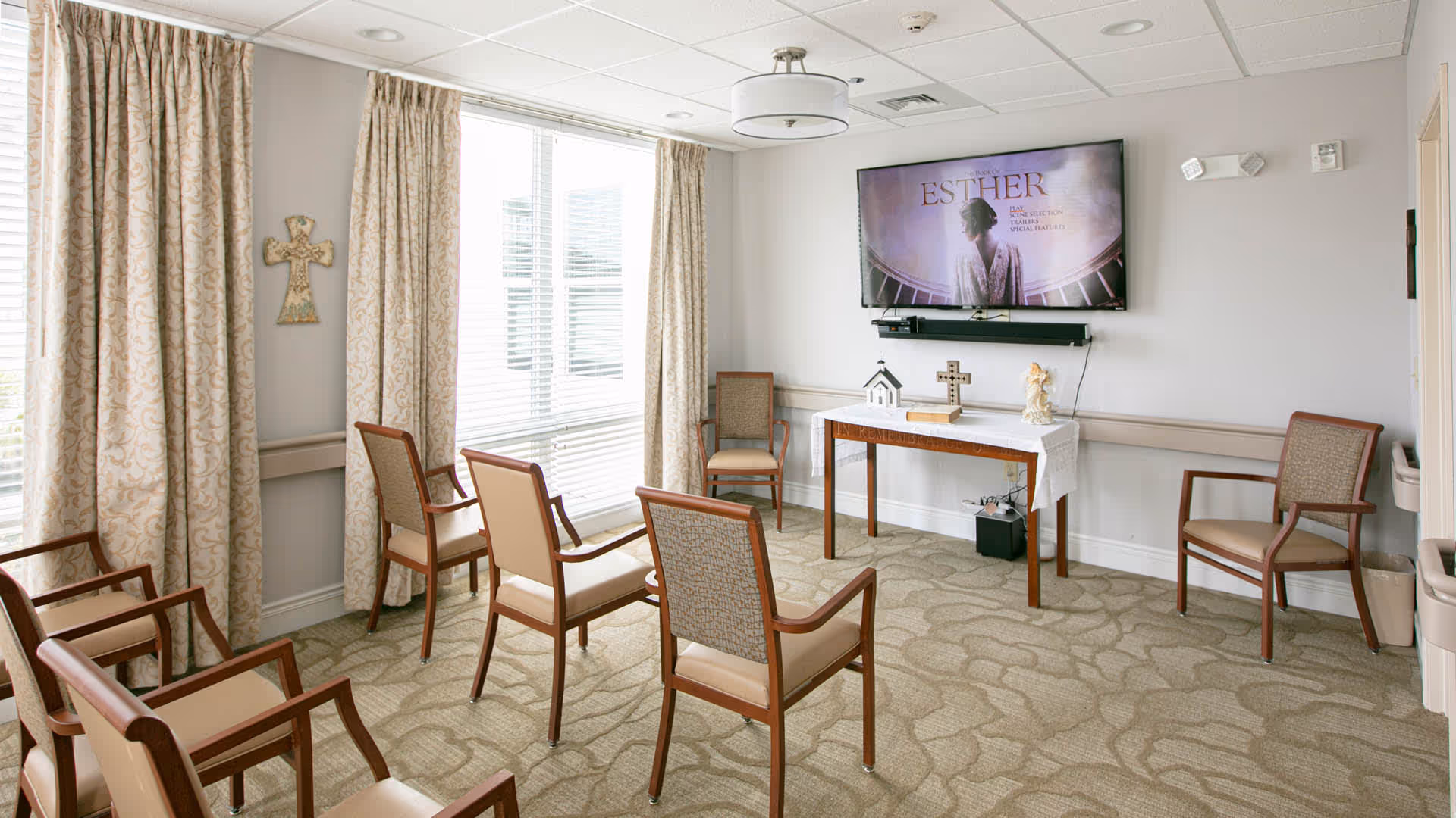 Small communal sitting room with chairs arranged facing a wall-mounted TV and a table displaying religious items.