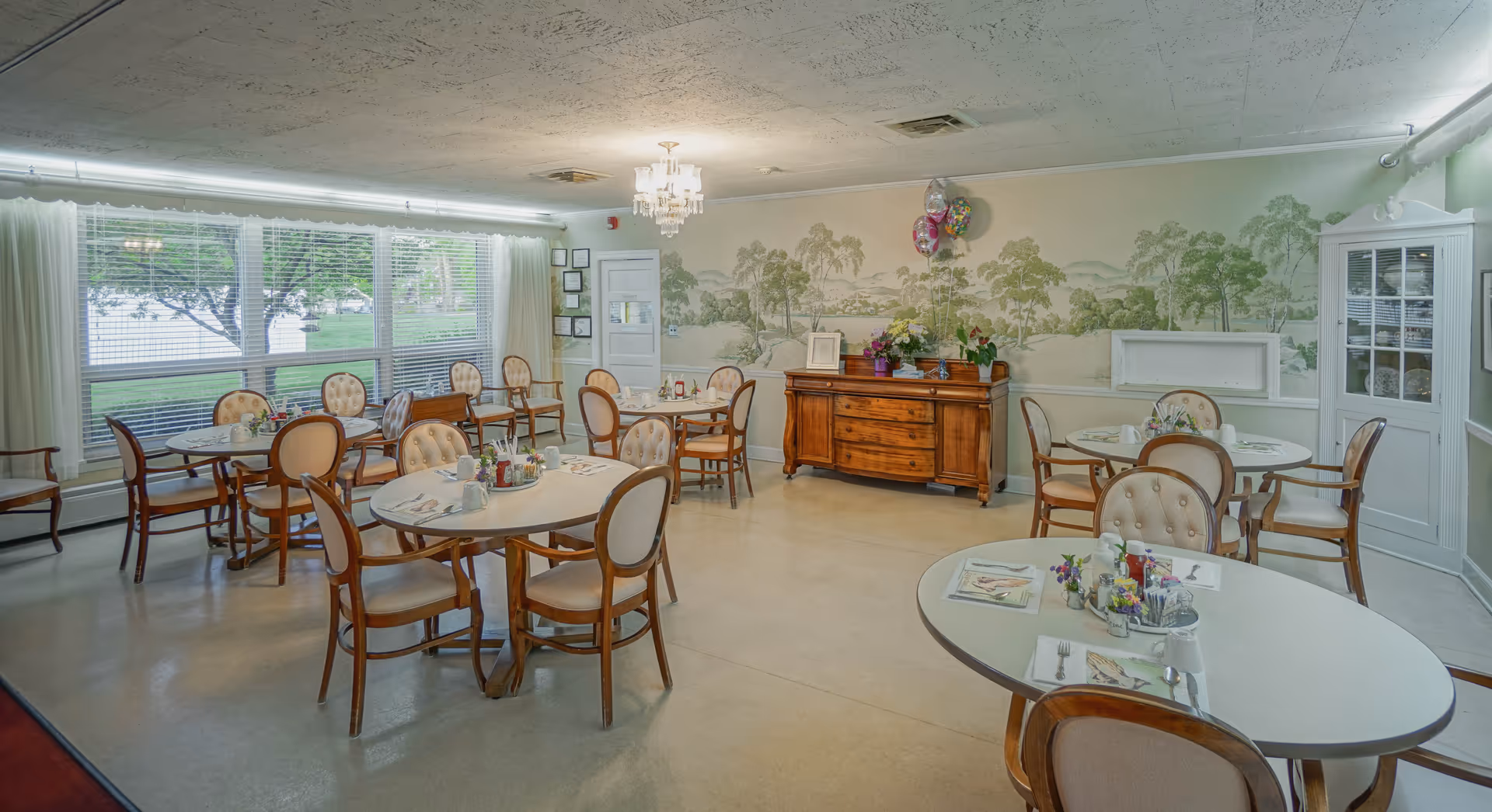 Bright dining room with round tables and upholstered chairs, place settings, and a wooden buffet against a muraled wall.