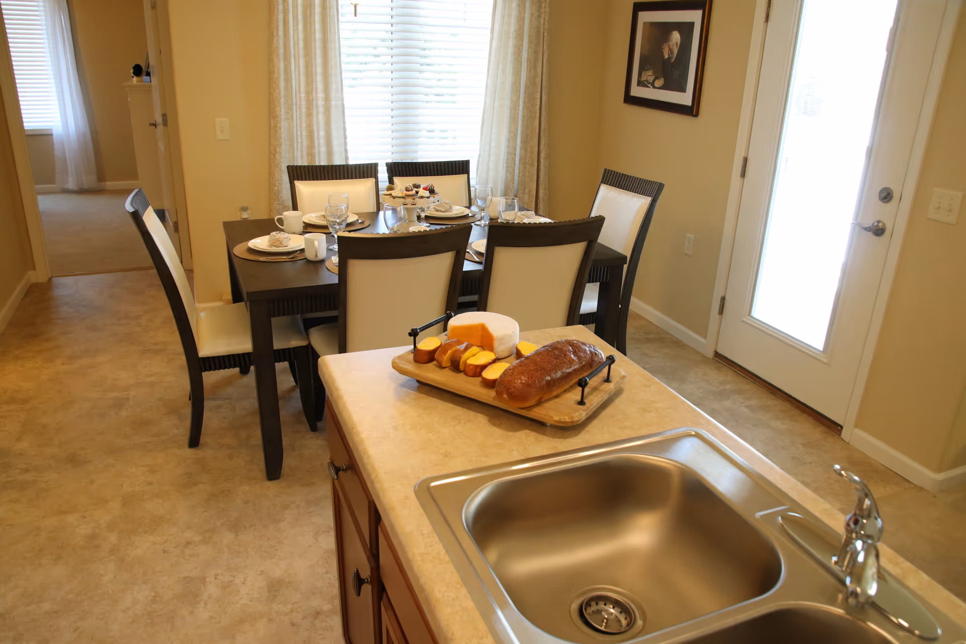 View of a dining area with a dark wooden table set for six people with plates, glasses, and cups. In the foreground, there is a kitchen island with a stainless steel sink and a wooden tray holding bread, cheese, and sliced fruit. The room has beige walls, a window with white blinds and curtains, and a door with a glass panel. A framed picture hangs on the wall near the door.