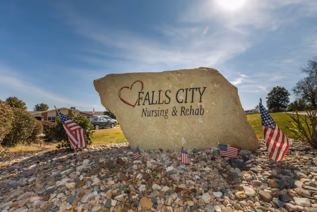 Large stone sign with a heart symbol and the text 'Falls City Nursing & Rehab' surrounded by small American flags and rocks, with a building and cars visible in the background under a partly cloudy sky.