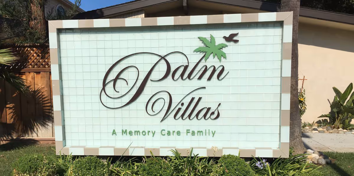 Outdoor sign for Palm Villas Memory Care facility with a decorative palm tree and bird graphic, surrounded by greenery and a wooden fence in the background.